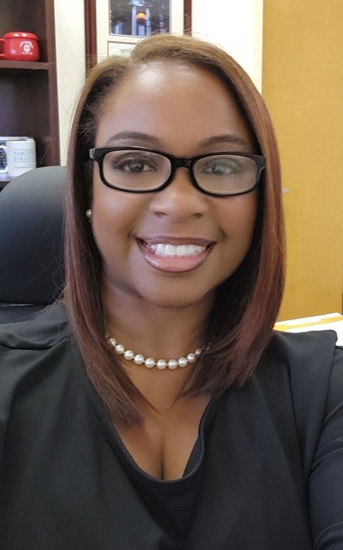 Woman with glasses and pearl necklace smiles, sitting at a desk.